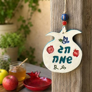 May include: A cream-colored ceramic pomegranate wall hanging featuring blue Hebrew text, a small blue bird, and two red pomegranate illustrations. The ornament is suspended by a jute string adorned with a blue and a red bead, hanging from a wooden post.