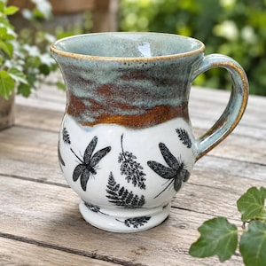 May include: A ceramic mug featuring a white base with black stamped dragonfly and fern leaf patterns. The upper portion of the mug has a glazed finish in shades of blue, green, and brown. It includes a curved handle and a flared rim design.