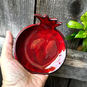 Peut inclure: Un bol en céramique rouge vif en forme de grenade avec une finition brillante. Le bol a un bord foncé et une surface texturée, tenu contre un fond en bois vieilli. Le bol mesure environ 13 cm de diamètre.