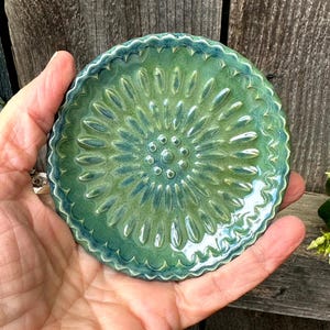 May include: A handmade, ceramic coaster with a floral design. The coaster is round, with a scalloped edge and a raised, textured pattern in shades of green and blue. The coaster is approximately 4 inches in diameter.
