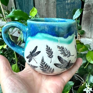 May include: A ceramic mug with a rounded body and a curved handle. The upper portion features a vibrant blue and turquoise glaze, while the lower section is off-white, decorated with black stamped illustrations of dragonflies and fern leaves.