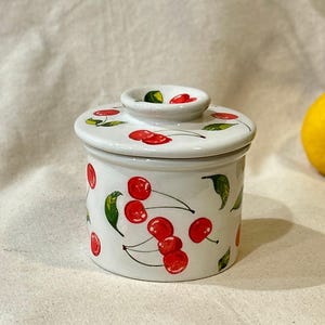May include: A white ceramic butter dish with a lid, decorated with red cherries and green leaves. The dish has a round shape and a small knob on the lid. A yellow lemon is visible in the background.