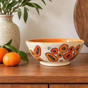 May include: A ceramic bowl with a speckled cream base and a bright orange interior. The exterior features hand-painted orange and black abstract papaya-like shapes with small black dots scattered across the surface.