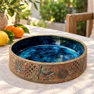 May include: A round ceramic bowl with a deep blue glazed interior and a textured, copper-toned exterior. The outer wall features intricate relief carvings of birds, dragonflies, and spiral patterns, interspersed with a decorative lattice design.