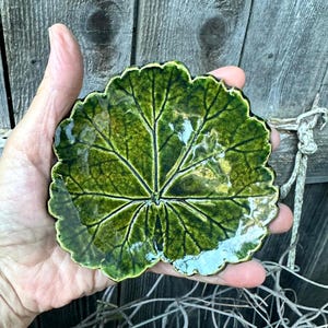 May include: A small, round, ceramic dish shaped like a green leaf with a glossy finish. The surface features detailed, dark green embossed veins that radiate from the center toward the scalloped, wavy edges of the leaf-shaped pottery piece.