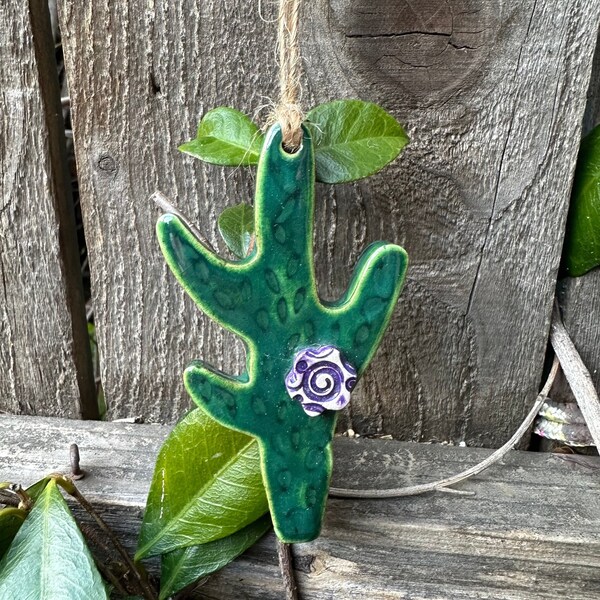 handcrafted ceramic cactus ornament, glazed in dark green, adorned with a lovely purple flower, suspended from a rustic hemp cord