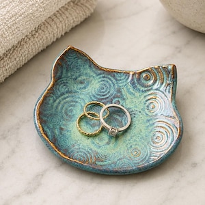 May include: A cat-shaped ceramic trinket dish with a teal and green glaze featuring a spiral pattern and gold-toned edges. Three metal rings, two gold-colored and one silver-colored, are resting in the center of the small, shallow dish.