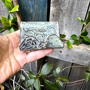 May include: A ceramic soap dish with a light green and brown glaze. The dish features a swirling cloud design on the top and a mountain-like shape on the front. The dish is approximately 4 inches wide.