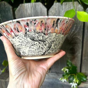 May include: A ceramic bowl with a unique design. The rim features a blend of red, orange, and black hues, while the body showcases a black and white illustration of birds and foliage. The bowl is held in a hand.