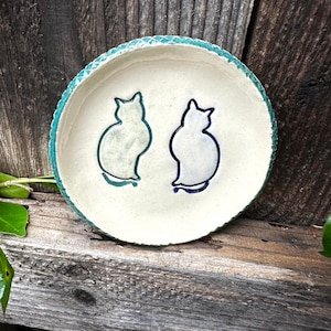 Puede incluir: Un plato de cerámica hecho a mano con un centro color crema y un borde turquesa. Dos siluetas de gatos están pintadas en el plato en tonos azules y verdes. El plato está sobre un fondo de madera rústica.