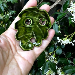 May include: A handcrafted, glossy green ceramic leaf with three circular holes. The leaf-shaped object has detailed vein lines and a smooth, reflective surface. The background features green foliage and white flowers.