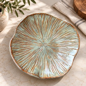 May include: A round ceramic plate with an irregular, wavy edge. The surface features a light seafoam green glaze with a radiating pattern of deep brown, textured lines that extend from the center toward the outer rim of the dish.