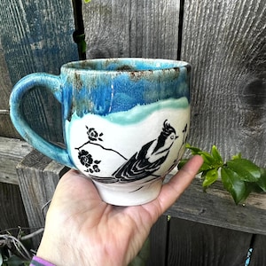Puede incluir: Una taza de cerámica hecha a mano con un esmalte azul y blanco. La taza presenta una ilustración de un pájaro en blanco y negro posado en una rama con flores. El asa es azul.