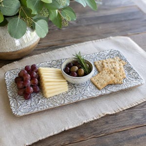 May include: A rectangular ceramic serving tray with a white and gray embossed floral pattern. The tray holds a cluster of red grapes, a stack of sliced cheese, a small round bowl containing olives and a rosemary sprig, and square crackers.