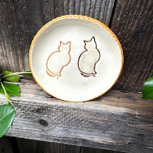Puede incluir: Un plato pequeño y redondo de cerámica con interior crema y borde texturizado marrón anaranjado. Dos siluetas de gato dibujadas en el interior, una en marrón claro y otra en marrón oscuro. Colocado sobre una superficie de madera envejecida.