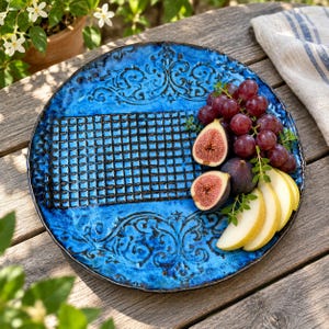 Puede incluir: Un plato redondo de cerámica azul vibrante con un patrón de rejilla oscuro en relieve en el centro y volutas ornamentales en los bordes. El plato está cubierto con un racimo de uvas moradas frescas, dos higos cortados y rodajas de pera.
