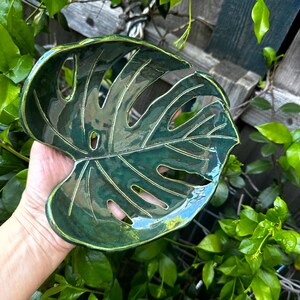 May include: A glossy, dark green ceramic dish shaped like a monstera leaf. The dish features realistic cutouts and embossed vein details throughout the surface, with a smooth, reflective finish and a slightly curved, organic edge profile.