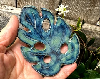 May include: A ceramic leaf-shaped decorative piece with a glossy finish. The leaf is a mix of blue and green hues, with three circular cutouts. The edges are outlined in silver. The piece is held in a hand, with a wooden background and greenery.