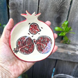 Peut inclure: Un plat en céramique en forme de grenade avec un bord rouge et un intérieur crème. Le plat présente des illustrations de fruits de grenade peintes à la main en rouge et noir. Le plat est tenu dans une main.