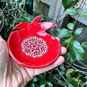 Peut inclure: Un plat en céramique rouge vif en forme de grenade, doté d'une finition brillante et d'un motif mandala blanc en relief au centre. Le plat présente une section supérieure pointue distincte rappelant la couronne d'une grenade.