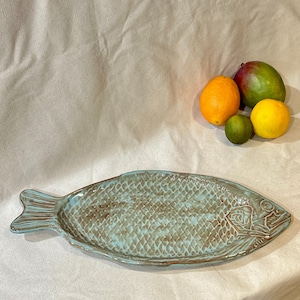 Puede incluir: Una fuente de cerámica turquesa con forma de pez, con un patrón de escamas texturizadas y un diseño detallado de cabeza de pez. La fuente está acompañada de una naranja, una lima, un limón amarillo y un mango.