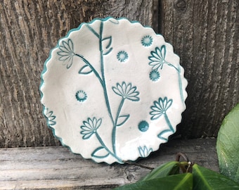 May include: A small, white ceramic dish with a scalloped edge. The dish is decorated with a teal blue floral design.