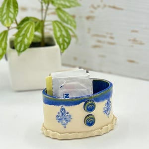 Op de afbeelding: Een witte keramische schaal met blauwe en groene accenten. De schaal heeft een gegolfde rand en een bloemenpatroon. Hij is gevuld met kleine witte pakjes.