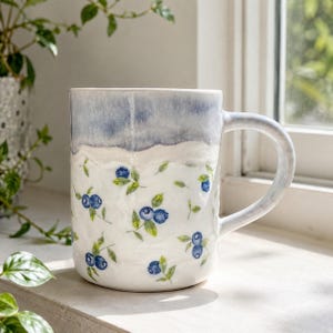 Pode incluir: Uma caneca de cerâmica com base branca apresentando um padrão pintado à mão de mirtilos azuis e folhas verdes. A parte superior da caneca tem um esmalte azul suave e mesclado que transita para o corpo branco. Inclui uma alça lateral.