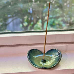 May include: A heart-shaped, ceramic incense holder with a glossy, teal and green finish. An incense stick is inserted into the holder, which sits on a white windowsill. The background shows a blurred view of green foliage.