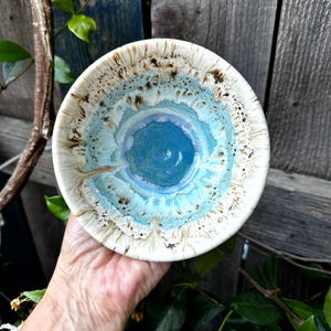 Könnte beinhalten: Eine handgefertigte Keramikschale mit einem einzigartigen Design. Die Schale hat einen tiefblauen Mittelpunkt, umgeben von konzentrischen Ringen aus hellblau, weiß und braun. Der Rand hat ein strukturiertes, gesprenkeltes Aussehen. Die Schale wird in einer Hand gehalten.
