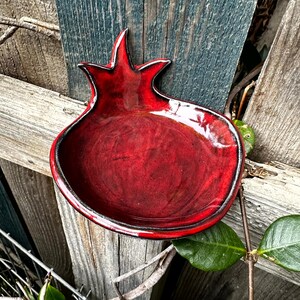Peut inclure: Un plat en céramique rouge fait à la main en forme de grenade, avec une finition brillante et une bordure noire. Le plat est posé contre une clôture en bois vieilli, mettant en valeur son design unique et sa couleur vibrante. Idéal pour servir ou exposer.