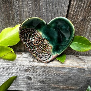 Puede incluir: Un plato de cerámica en forma de corazón con un esmalte verde y un diseño de encaje marrón. El plato está sobre una superficie de madera.