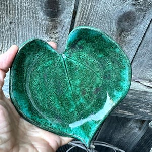 May include: A heart-shaped ceramic dish featuring a vibrant emerald green glaze with a mottled, textured finish. The surface displays a subtle embossed vein pattern resembling a leaf, with darker green and black accents throughout the glaze.