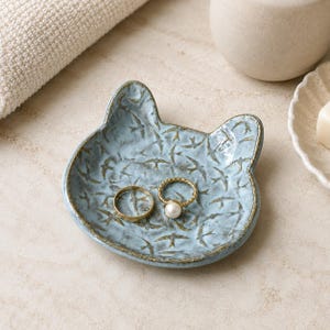 May include: A light blue ceramic trinket dish shaped like a cat head, featuring a textured surface with a repeating pattern of small dark birds. Two gold-toned rings, one plain and one with a white pearl, rest inside the dish.