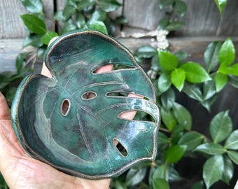 May include: A green ceramic soap dish shaped like a monstera leaf. The dish has a glossy finish and a textured surface.