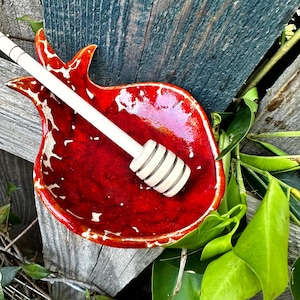 Peut inclure: Un plat en céramique rouge en forme de grenade avec un bâtonnet à miel blanc. Le plat a une finition brillante avec des accents blancs. Le bâtonnet à miel est en bois de couleur claire. Les articles sont posés sur un fond en bois vieilli.