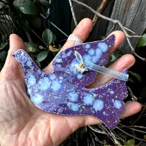 May include: A ceramic dove ornament in shades of purple and blue, speckled with lighter blue and yellow accents. The dove is held in a hand, and has a ribbon for hanging. A small gold charm and a light blue ribbon are attached.