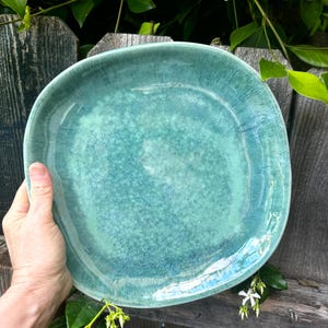 Puede incluir: Plato de cerámica turquesa hecho a mano, con una forma irregular. El plato tiene una superficie texturizada y un acabado brillante, con variaciones de color. El plato está sostenido por una mano, mostrando su tamaño y diseño.