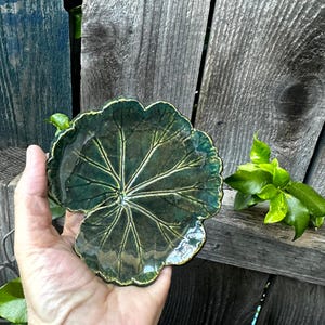May include: A handcrafted, leaf-shaped ceramic dish with a glossy, dark green glaze. The dish features detailed veining and a slightly raised edge. The unique design is reminiscent of nature and adds a touch of organic elegance to any space.
