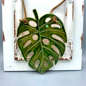 tropical green monstera leaf crafted in ceramic, suspended elegantly by a leather cord, botanical art piece, bring nature's beauty indoors