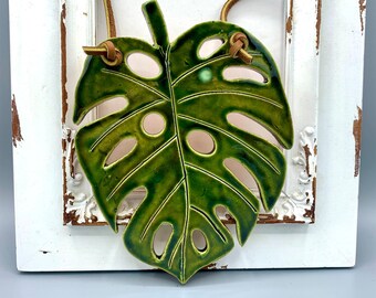 tropical green monstera leaf crafted in ceramic, suspended elegantly by a leather cord, botanical art piece, bring nature's beauty indoors