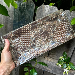 Puede incluir: Una fuente rectangular de cerámica con un detallado patrón floral y geométrico en tonos marrón, crema y verde azulado. El plato hecho a mano tiene un acabado brillante y se sostiene contra un fondo de valla de madera.