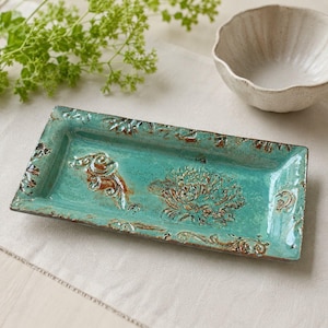 May include: A rectangular ceramic tray with a glossy turquoise glaze and distressed brown edges. The surface features embossed floral and scrollwork patterns. The tray has a raised rim and a textured finish with visible variations in color.