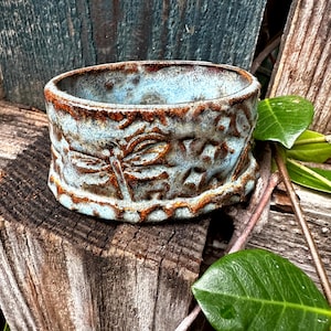 May include: A handcrafted ceramic bowl with a blue and brown glaze. The bowl features a dragonfly design and other embossed patterns around the exterior. The bowl is sitting on a wooden surface.