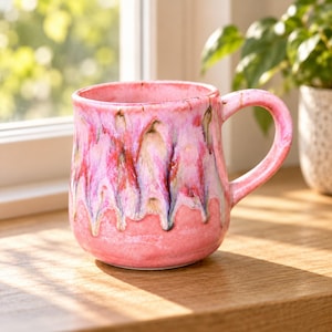 May include: A ceramic mug with a rounded body and a curved handle. The mug features a pink base glaze with a decorative drip pattern in shades of magenta, purple, and white cascading from the rim down toward the center of the vessel.