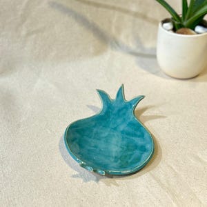 Peut inclure: Un plat en céramique turquoise en forme de grenade. Le plat a une surface texturée et un bord surélevé. Le design de la grenade comprend une tige et une base. Le plat est sur une surface de couleur neutre, avec un petit pot blanc avec une plante en arrière-plan.
