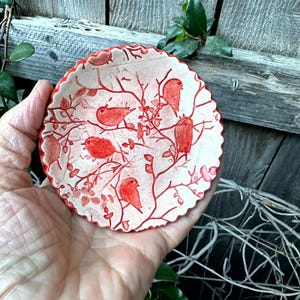May include: A small, round ceramic dish with a scalloped edge, featuring a cream-colored base and a detailed red pattern of multiple birds perched on thin, branching twigs. The design covers the entire interior surface of the shallow bowl.