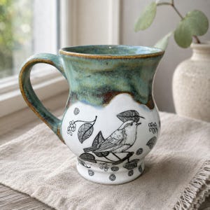 May include: A ceramic mug featuring a white lower section with a black line drawing of a bird perched on a branch surrounded by leaves and berries. The upper portion of the mug has a mottled teal and brown glaze with a curved handle on the side.