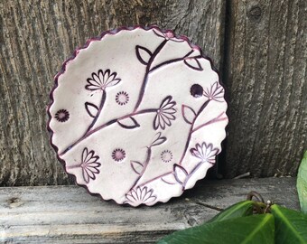 May include: A small white ceramic dish with a scalloped edge and a floral design in shades of burgundy. The dish features stylized flowers and leaves.