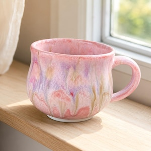 May include: A ceramic mug with a rounded body and a curved handle, featuring a glossy glaze in shades of pastel pink, soft lavender, and muted tan. The colors blend in a vertical drip pattern across the exterior surface, ending at a white base.